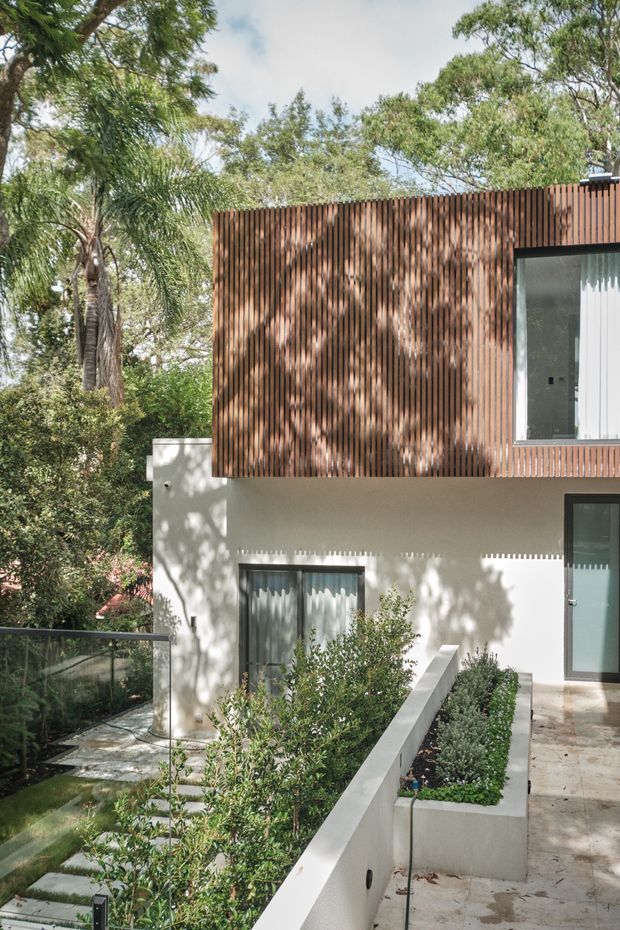 Exterior view of a contemporary multi-level residence set among mature trees and landscaped planting. The facade combines smooth light-rendered walls with a prominent upper-storey screen of closely spaced vertical timber battens, dark-framed glazing, and a paved terrace with raised garden beds and a glass balustrade.