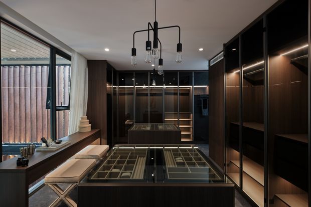 A dark, sophisticated walk-in wardrobe or dressing room with extensive floor-to-ceiling dark timber cabinetry, black-framed glass doors, illuminated shelving and drawers, and a central island with glass-topped display compartments. A narrow window provides natural light, while a sculptural black pendant light hangs centrally above the island. Two upholstered stools sit along the window-side counter.