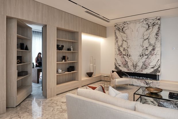 Luxurious contemporary living room with pale timber built-in shelving and cabinetry, polished marble flooring, a large dramatic veined marble feature wall surrounding a recessed fireplace, and light upholstered seating. A glazed or mirrored opening reveals an adjacent area, while integrated linear ceiling ventilation and lighting reinforce the refined minimalist fit-out.