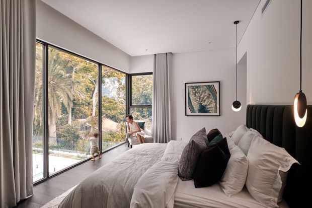 A refined contemporary bedroom with a large upholstered bed, layered neutral bedding and cushions, and a black channel-tufted headboard. Full-height corner glazing with curtains opens the room to leafy greenery, while sculptural pendant lights and framed artwork add understated visual interest.