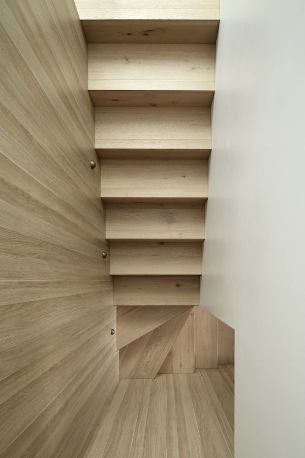A narrow, minimalist stairway viewed from above, with light oak timber treads, matching timber wall panelling and flooring, and crisp white walls. The staircase includes a compact turn with angular timber detailing and concealed-looking construction, creating a warm, streamlined architectural interior.
