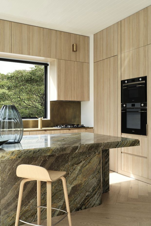 A refined contemporary kitchen featuring full-height light timber cabinetry, integrated black double ovens, a gas cooktop, and a substantial sculptural island clad in richly veined dark natural stone. Pale timber flooring and a white-lined ceiling create a warm, cohesive palette, while a large window brings in greenery and daylight. A minimalist timber bar stool and decorative glass vessel sit at the island.