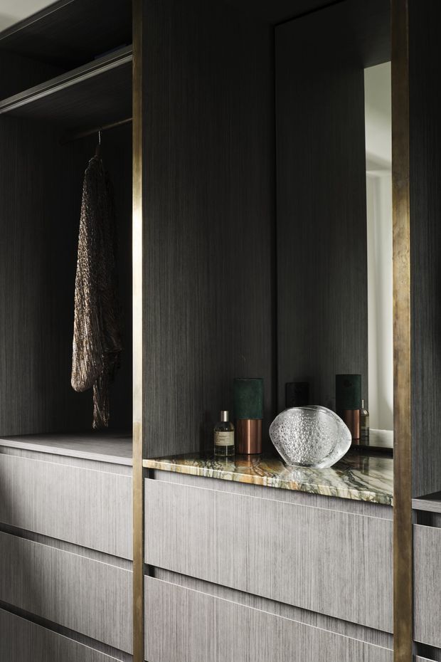Close-up of a refined walk-in wardrobe or dressing-room joinery installation. Dark vertically textured cabinetry frames open hanging storage and pale grey drawers, with a marble or richly veined stone benchtop, aged brass-toned vertical trim, and a full-height mirror. Small accessories sit on the counter, creating a moody, luxurious, highly detailed interior.