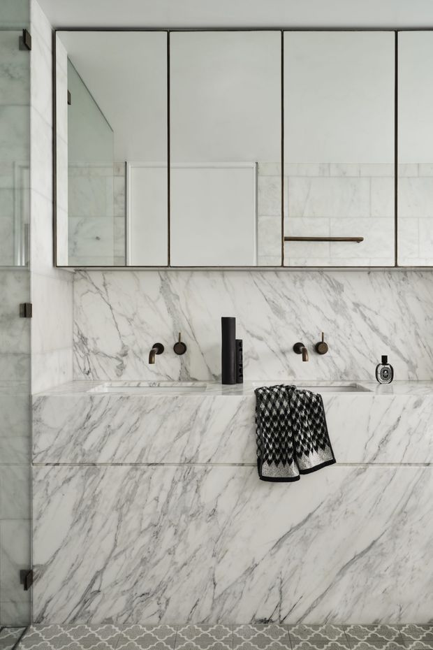 A refined contemporary bathroom vanity featuring a double basin carved into a white marble-clad counter and wall. The space includes mirrored upper cabinetry with slim dark frames, wall-mounted bronze-toned taps, a matte black central mixer, and patterned grey floor tiles. A black-and-white towel adds contrast.