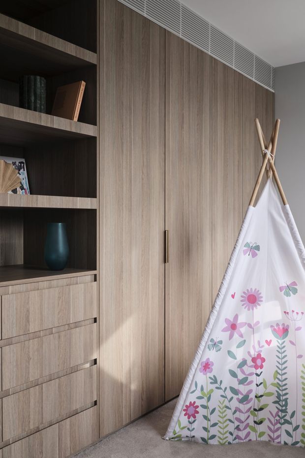 A refined children's bedroom storage wall featuring full-height light timber wardrobe doors, integrated open shelving and drawers, a linear ventilation grille near the ceiling, and a small floral-patterned teepee in the foreground. The joinery has a warm, contemporary appearance with dark recessed shelving and understated hardware.