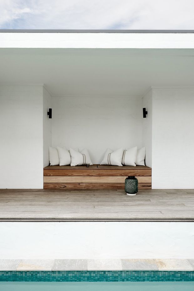 Minimalist poolside outdoor lounge area with a built-in timber daybed recessed into a white wall, lined with white cushions. Pale timber decking leads to a turquoise tiled swimming pool, with simple black wall lights and a small lantern adding contrast.