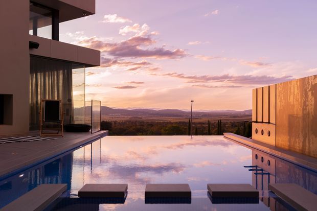 A contemporary modern residence viewed from the pool terrace at sunset. The scene features a reflective infinity-edge swimming pool with submerged stepping platforms, a cantilevered flat-roofed building with expansive glazing on the left, and a warm-toned vertical-panel privacy wall on the right. Rolling hills and a broad valley are visible beyond the property, while the sunset creates a soft pink, purple, and warm orange atmosphere.