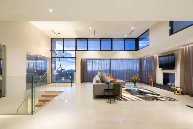 A spacious, contemporary open-plan living room with a polished light floor, high ceiling and extensive floor-to-ceiling and clerestory glazing. A grey sectional sofa faces a wall-mounted television and sleek linear fireplace, with a colourful patterned rug and sculptural pendant light adding visual interest. A glass balustrade and floating timber stair treads lead to a lower level, while sheer curtains soften the large windows.