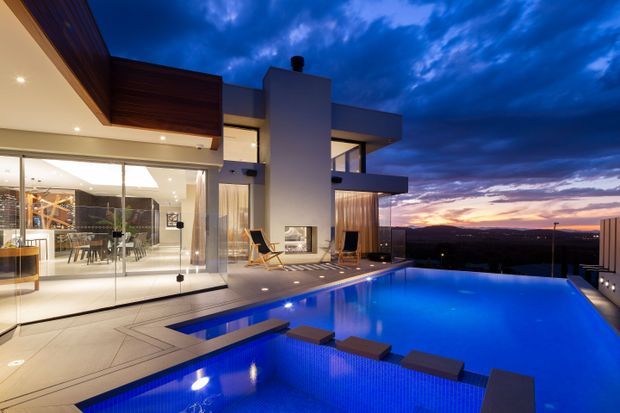 Twilight exterior view of a modern multi-level residence with extensive floor-to-ceiling glazing, projecting flat-roofed volumes, warm timber soffits, and a large illuminated swimming pool in the foreground. The home overlooks a distant landscape and sunset horizon, with an indoor dining and living area visible through the glass.