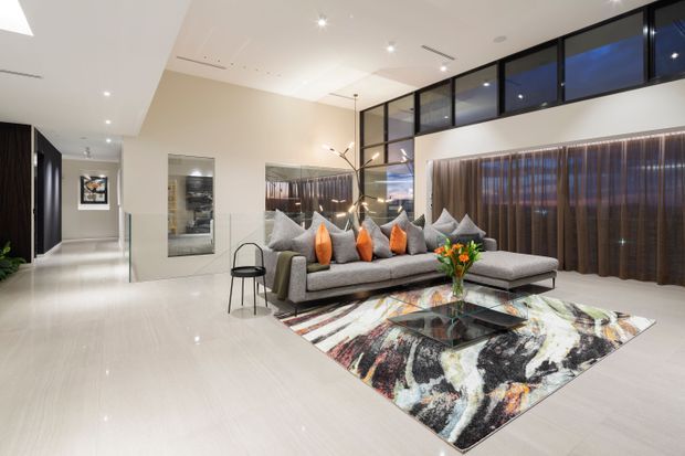 A spacious contemporary living room with a large grey L-shaped sectional sofa, vivid orange accent cushions, a glass coffee table, and an abstract patterned area rug. Full-height dark-framed glazing and sheer curtains line the rear wall, while recessed ceiling lights, pale flooring, and a double-height feel create a bright, polished interior. A hallway and adjoining spaces are visible to the left.