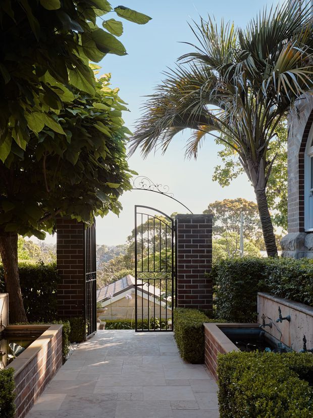 A landscaped residential garden entry viewed from a paved path, featuring an ornate black metal pedestrian gate between dark brick pillars, clipped hedges, mature broadleaf planting and a palm tree. A descending view toward neighbouring rooftops and a bright suburban landscape is visible beyond the gate. The house edge at right has light masonry and dark-framed glazing.