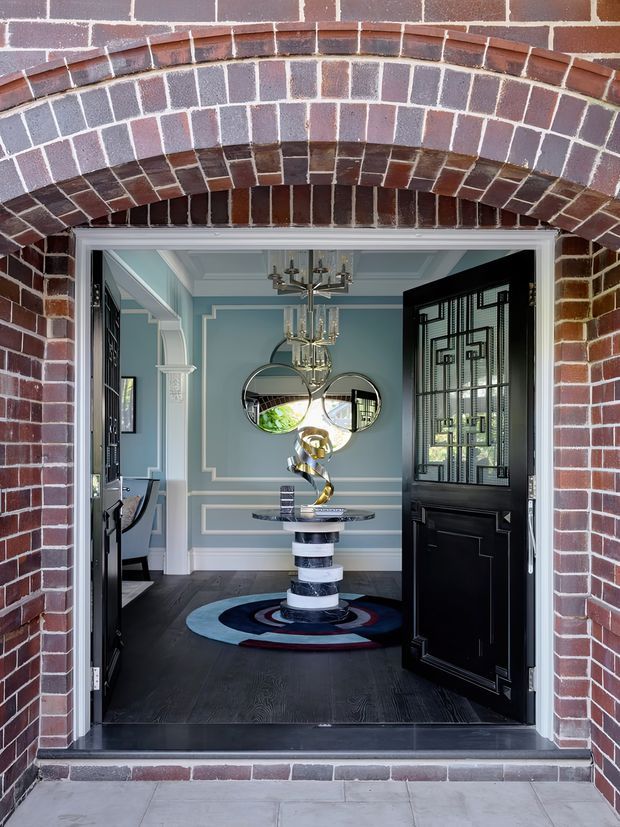 View through an arched red-brick exterior entry into a refined foyer. The interior features pale blue-grey walls with white decorative moulding, dark timber flooring, black glazed-panel doors, a sculptural central console, circular wall mirrors, a crystal chandelier, and a bold round patterned rug. The entry is framed by an ornate brick arch and opens directly into the home.