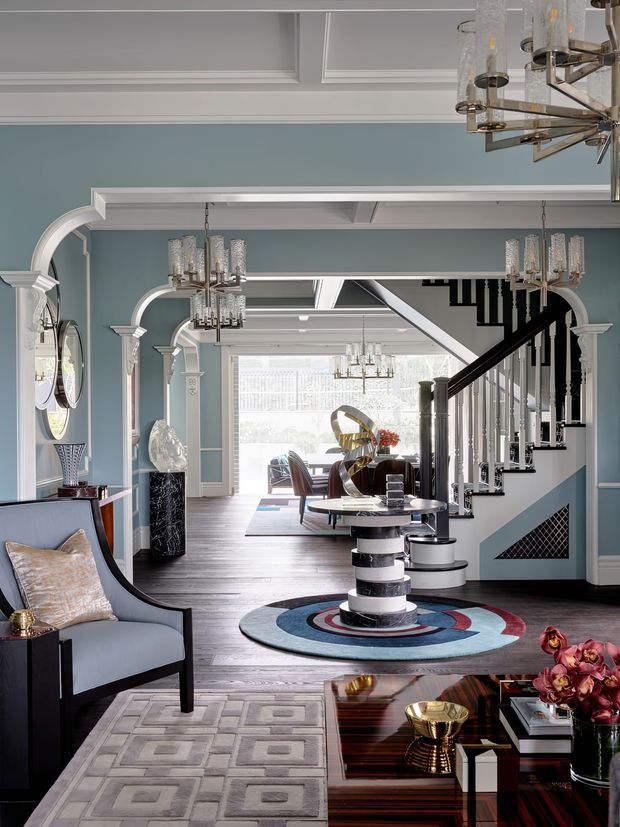 A refined, highly styled residential living room and entry hall with pale blue walls, extensive white architectural trim, repeated arched openings, dark timber flooring, and a dramatic black-and-white staircase. The room features a sculptural circular rug, modern lounge seating, a glossy dark coffee table, decorative objects, mirrored wall accents, and multiple crystal pendant chandeliers. Bright glazing at the rear opens the space visually toward a dining or living area.