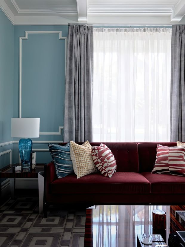 A refined living room with a deep burgundy velvet sofa, patterned accent cushions, a glossy black side table, and a blue glass table lamp. The room features blue-green painted walls with white decorative wall moulding, an ornate coffered ceiling, full-height windows with sheer white curtains and grey drapes, and a geometric patterned rug. The overall composition is polished, colourful, and classically detailed.