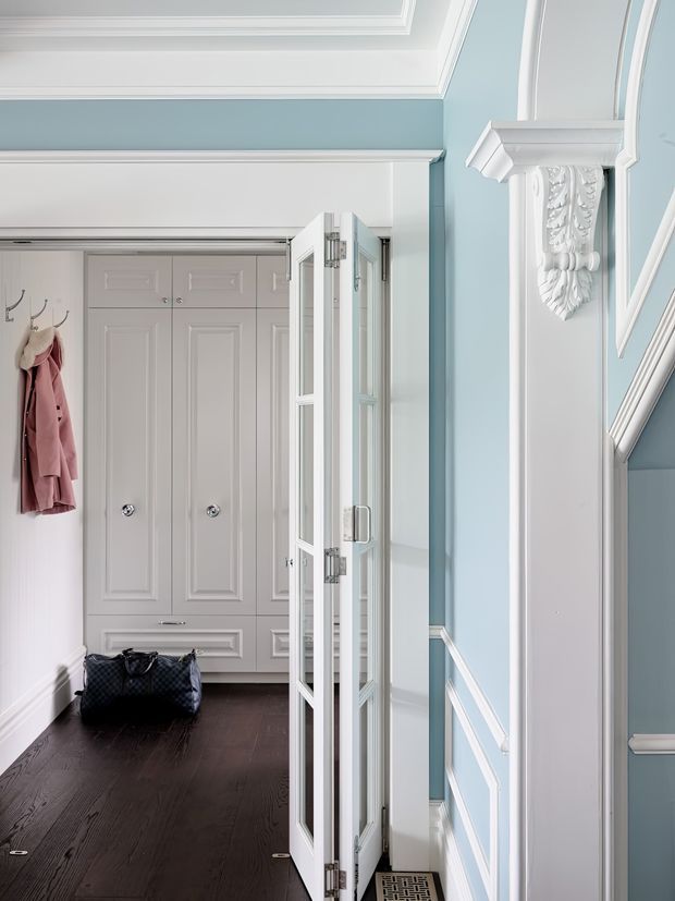 A refined residential entry hall with pale blue walls, white decorative wall panelling and ornate moulding, a dark timber floor, and tall built-in white cabinetry at the far end. A glazed white-framed door stands open beside the hall, revealing a stairway edge on the right. Wall hooks with a coat and a dark travel bag add practical entryway details.