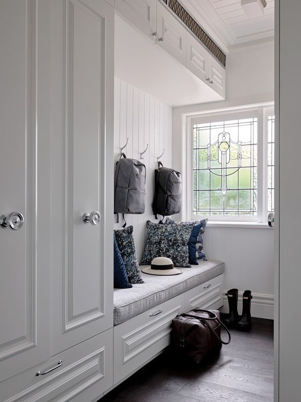 A bright, traditionally detailed mudroom or entryway with extensive white built-in cabinetry, paneled doors, an integrated upholstered window seat with drawer storage, coat hooks, and a decorative leaded-glass window. Patterned blue and grey cushions, bags, a hat, and boots add practical entry storage and a lived-in feel. The room has dark timber flooring, white wall panelling, ornate trim, and a coffered or panelled ceiling.