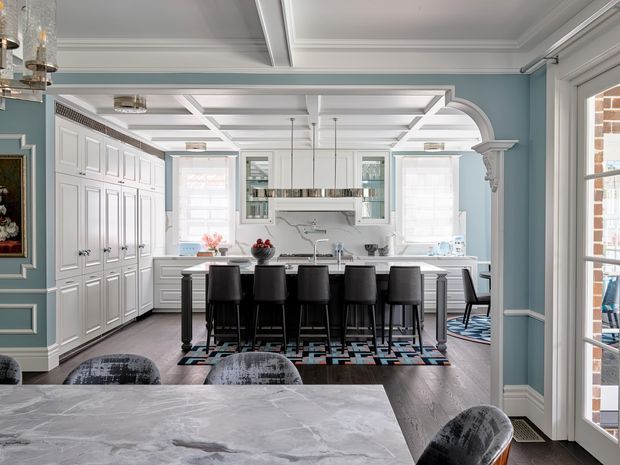 A refined, spacious kitchen and dining area with pale blue walls, extensive white built-in cabinetry, a large central island with seating for six, and a polished light stone benchtop. The room features elaborate coffered ceilings, detailed white mouldings, an arched opening with a decorative column, pendant lighting, dark timber flooring, and a patterned area rug. French doors and windows bring in abundant natural light.