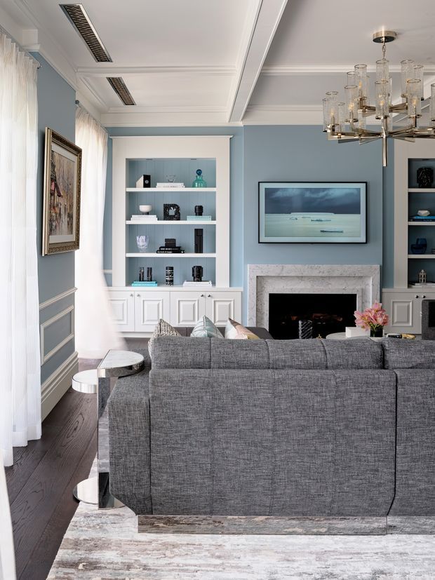 Refined living room viewed from behind a large grey sectional sofa. The room features pale blue walls with white decorative wainscoting and built-in shelving, dark timber flooring, a marble fireplace surround, a wall-mounted artwork or television, and a statement glass-and-metal chandelier. Tall sheer curtains provide soft natural light, while curated shelving and classic trim details create an elegant, tailored interior.