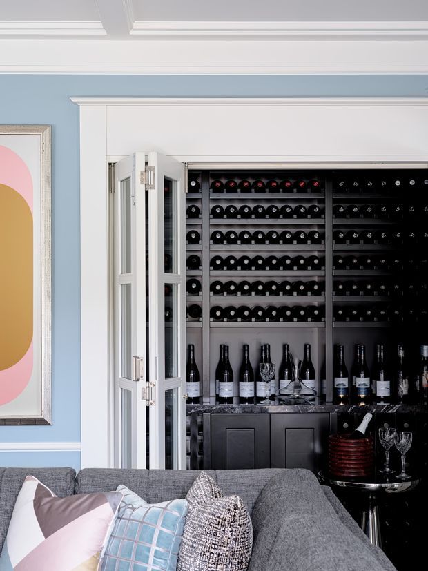 A refined living room corner with a large grey sectional sofa in the foreground and a built-in, glass-fronted wine storage cabinet behind it. The wine display includes extensive bottle racks, lower cabinetry, bottles, glassware and a small serving area. The room has pale blue walls, white decorative wall panelling and ornate coffered ceiling mouldings, with a colourful abstract artwork on the wall. The overall look is elegant and luxurious with a strong entertaining focus.