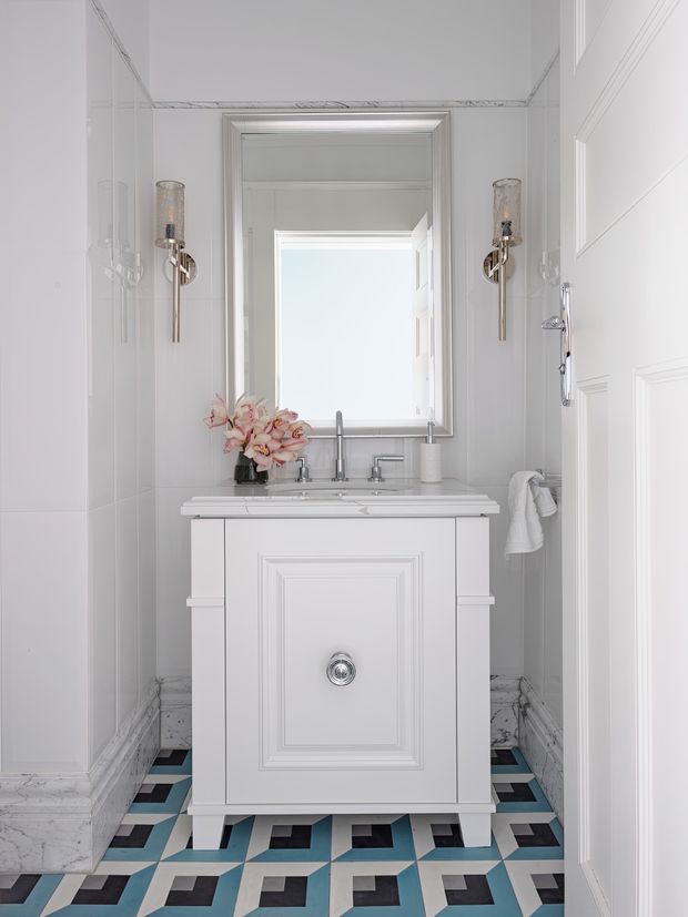 A compact, refined bathroom featuring a white paneled vanity with a marble countertop, twin chrome faucets, a large framed mirror, and wall-mounted brass sconces. The room has white paneled walls and trim, marble-look lower wall detailing, and a bold geometric floor in white, black, and turquoise-blue. A towel hangs beside the vanity and a small floral arrangement adds a soft decorative accent.