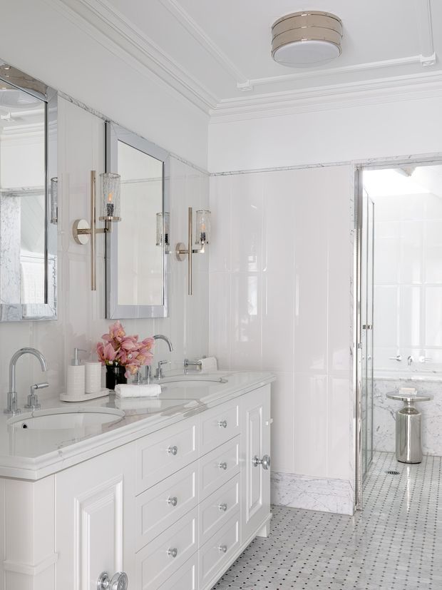 Bright, elegant white bathroom featuring a long double vanity with raised-panel cabinetry, two integrated basins, chrome fixtures, mirrored wall panels, decorative wall sconces, glossy white wall tiles, and a small-format patterned tile floor. A glass-panel opening leads to a shower or bathing area with matching white and marble-patterned finishes. The room has ornate ceiling mouldings and a refined classic aesthetic.