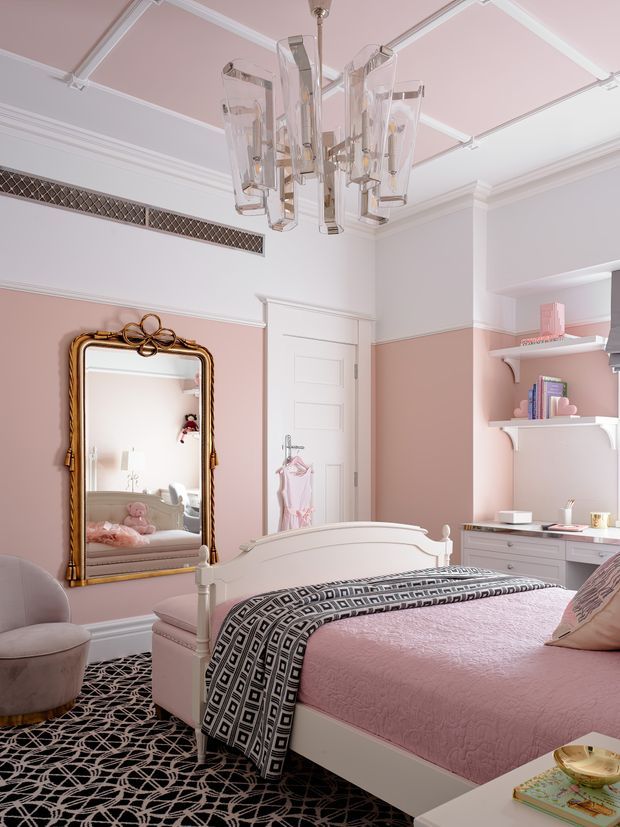 A refined pink-and-white bedroom with a traditional panelled door, built-in open shelving and desk, ornate gold-framed mirror, upholstered white bed, pink bedding, and a striking geometric black-and-white carpet. A sculptural glass chandelier hangs from a coffered ceiling with pink inset panels, creating a polished, feminine interior.