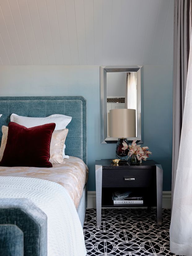 A refined bedroom corner with a blue upholstered bed and matching blue wall, layered white bedding, and a deep burgundy accent cushion. A black bedside table with a lamp, flowers, books, and a large framed mirror sits beside the bed. The room features white vertical wall panelling and a bold black-and-white geometric carpet, with sheer curtains at the window.