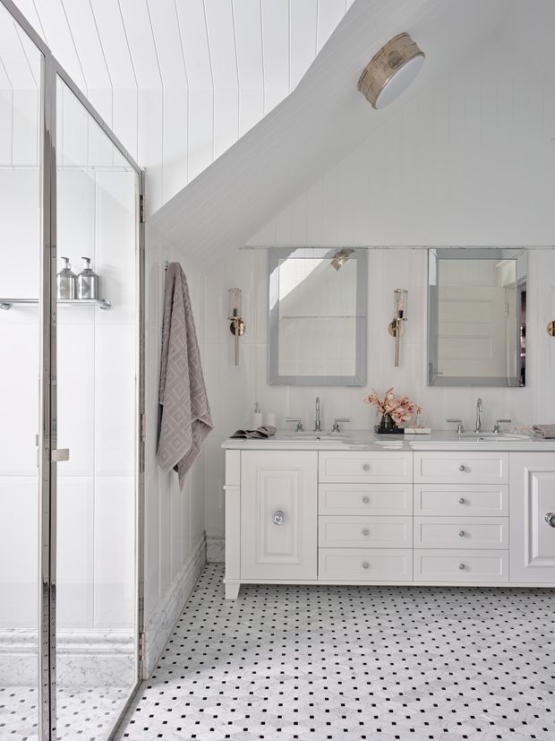 Bright white bathroom with a long traditional vanity featuring multiple drawers and inset-panel cabinetry, twin rectangular mirrors, polished metal wall sconces, and a glass-enclosed shower at left. The room has a sloped white paneled ceiling, white wall surfaces, and a white tiled floor patterned with small black square accents. A pale fabric towel hangs beside the shower, and the overall styling is crisp, classic, and refined.
