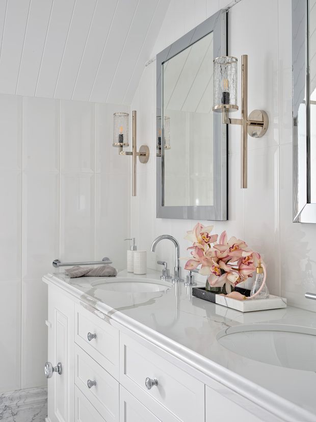 Bright, elegant white bathroom vanity with two integrated basins, paneled cabinetry, a pale stone countertop, polished chrome fixtures, a large framed mirror, and decorative wall sconces. Glossy white wall panels and a coffered or paneled ceiling create a refined, traditional feel, while orchids and small accessories add a soft decorative accent.