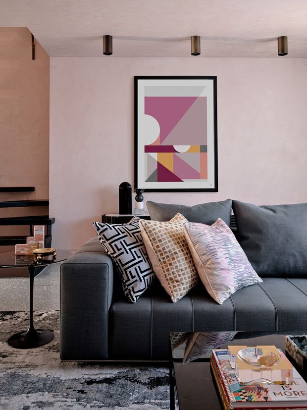 A styled contemporary living room featuring a large charcoal-grey sectional sofa with patterned cushions, a black glass coffee table, an abstract geometric artwork in a black frame, a textured grey rug, and dark open shelving. The room has soft blush-pink walls and ceiling with slim black and brass-toned track-style spotlights, creating a refined, moody interior.