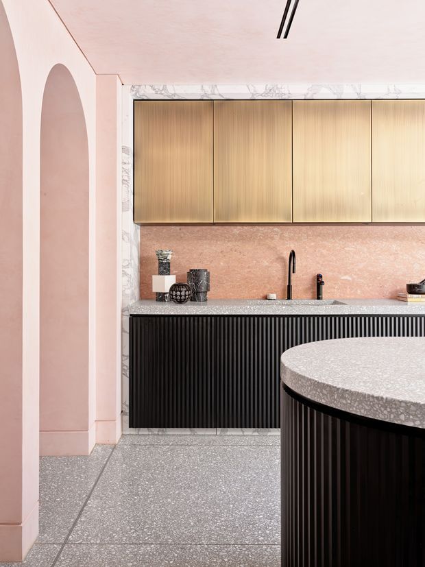 A refined contemporary kitchen featuring pale pink plaster walls with a tall arched niche, warm vertically grained timber overhead cabinetry, black ribbed base cabinetry, a pink stone backsplash, and terrazzo flooring. A large rounded island with a terrazzo benchtop and dark fluted cabinetry occupies the foreground. Marble-look trim frames the rear cabinetry, while slim black linear ceiling lights provide understated illumination.