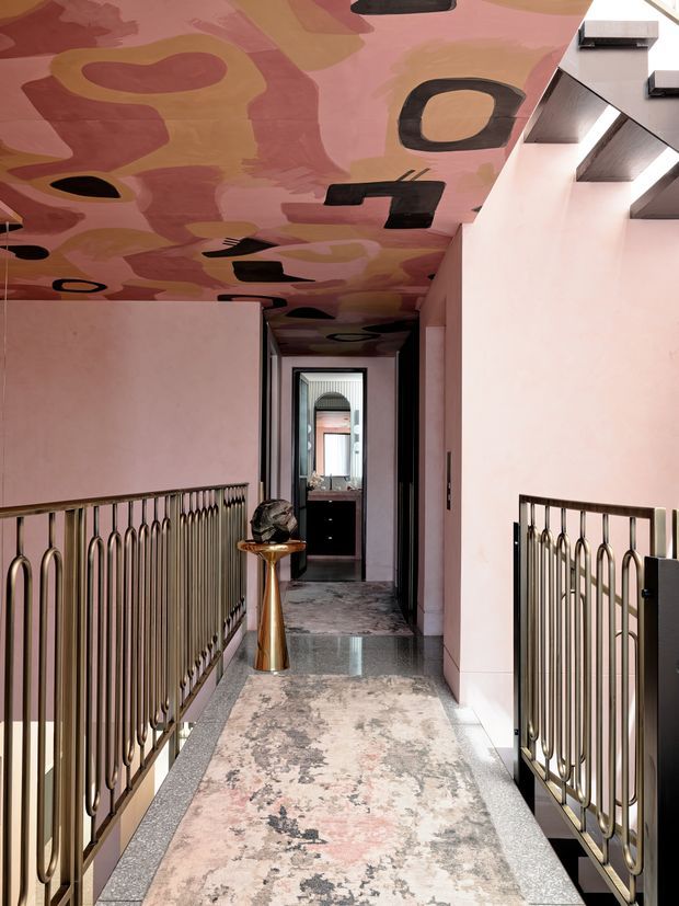 A refined residential hallway or landing with soft pink walls, an expressive painted ceiling featuring abstract organic forms in pink, terracotta, ochre and black, and ornate dark metallic railings with looping motifs. A patterned runner extends along the polished stone or terrazzo floor toward a doorway and dark built-in cabinetry. A small gold-toned pedestal table with sculptural decor adds a luxe accent. The space is narrow, sophisticated and highly decorative, with a strong view down the corridor.
