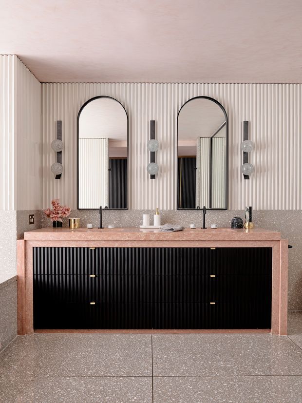 A refined contemporary bathroom vanity with a wide double-sink arrangement, arched black-framed mirrors, black fluted cabinetry, and a pink-toned stone benchtop. The walls feature vertical fluted panelling above a speckled stone dado, complemented by sculptural wall lights and terrazzo-style flooring.