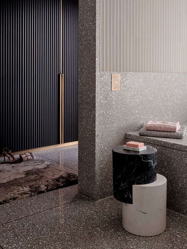 A refined contemporary interior detail showing a dark vertically ribbed wall or door with a slim brass-toned handle, terrazzo flooring and wall surfaces, and a sculptural black marble and white cylindrical side table. A raised terrazzo ledge holds folded textiles, while a textured dark rug and small decorative objects add contrast.