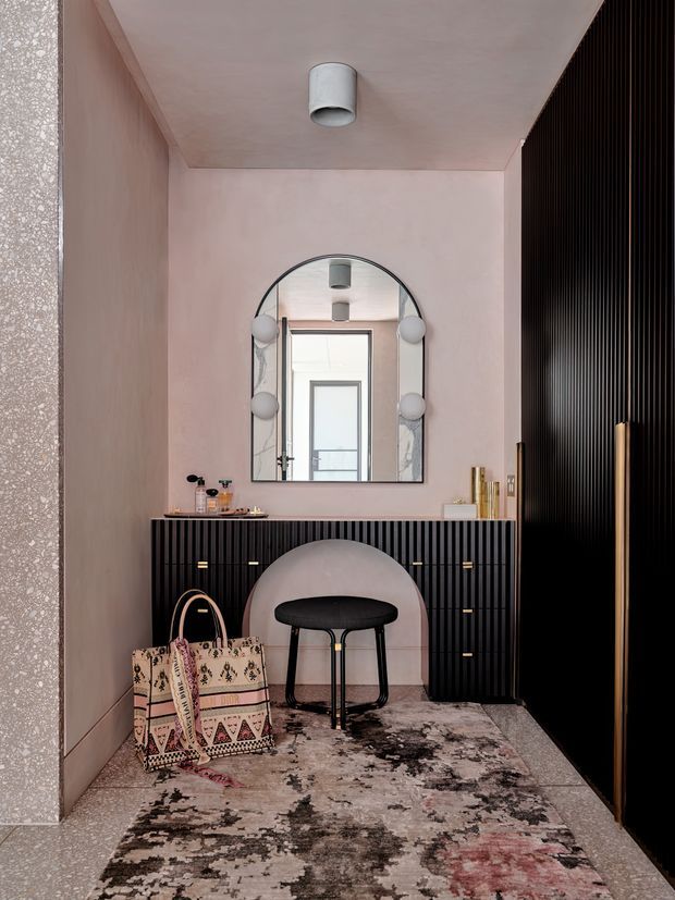 A refined interior dressing or entry area with blush pink plastered walls and ceiling, a black vertically fluted built-in console with brass pulls, and a matching dark fluted wall panel. An arched illuminated mirror with round side lights hangs above the console, accompanied by a sculptural upholstered stool, patterned area rug, decorative bag, and small accessories. A doorway and additional interior space are reflected in the mirror. The composition is compact, polished, and softly lit with a contemporary luxe character.