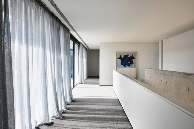 A bright contemporary interior corridor or landing with floor-to-ceiling glazing concealed behind sheer grey curtains on the left. A grey carpeted walkway leads past a white half-height partition and a raw concrete feature wall, with a framed abstract artwork adding a small visual accent. The space has clean lines, a minimalist palette, and concealed linear ceiling ventilation.
