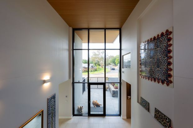 A tall, bright residential entryway viewed from a stair landing, featuring floor-to-ceiling black-framed glazing and a glazed door opening to a patio and front garden. The space has white walls, pale tiled flooring, a warm timber-lined ceiling, contemporary wall lighting, and decorative patterned textile or tile wall art. A staircase descends at the left, while built-in joinery is visible beside the entry door.