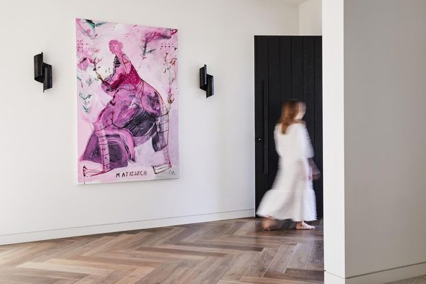 A bright, minimalist interior hallway or entry space with white walls, a large pink figurative artwork, black sculptural wall sconces, a dark vertical-panel door, and a patterned timber parquet floor. A blurred person in a white dress is moving through the doorway.
