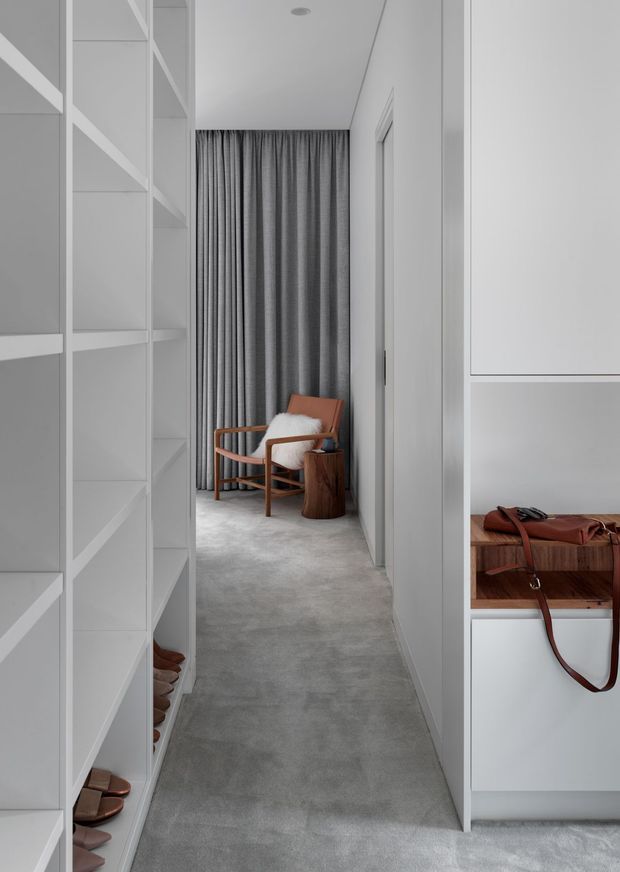 A narrow, highly organized walk-in wardrobe with floor-to-ceiling white built-in shelving and cabinetry on both sides. The space has a soft grey carpeted floor, a full-height grey curtain at the end, and a timber-framed lounge chair with a white cushion beside a small rustic timber stool. Open shelves display shoes, while a leather bag rests on a built-in bench or drawer unit. The overall design is restrained, monochromatic, and contemporary with warm timber and leather accents.