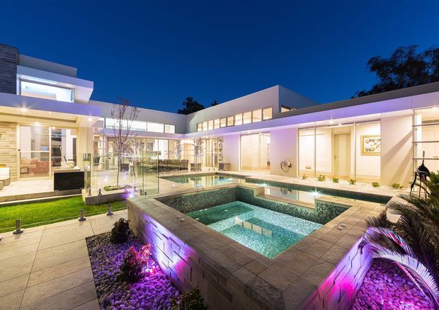 Twilight exterior view of a large contemporary flat-roof residence organised around a landscaped courtyard and multiple illuminated swimming pools. The scene features crisp white rendered forms, extensive floor-to-ceiling glazing, pale stone or concrete paving, a raised spa or pool edge, planted pebble beds, and colourful architectural lighting.