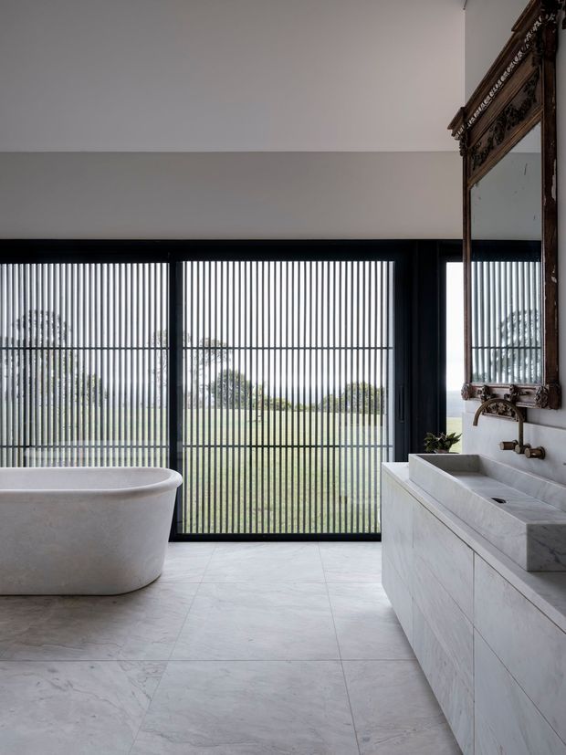 A refined contemporary bathroom with a freestanding white bathtub, double stone vanity, wall-mounted brass fixtures, and an ornate dark wood-framed mirror. Large black-framed glazed doors with vertical privacy screening overlook a landscaped outdoor area. The space is bright, restrained, and highly material-focused, with pale stone surfaces and strong black framing.