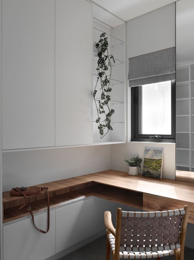 A compact, carefully designed home office nook with an L-shaped timber desk integrated into white built-in cabinetry. The space includes a black-framed window with a grey roller blind, open shelving, a trailing indoor plant, artwork, and a woven leather-and-timber chair. The palette is restrained and contemporary, with warm timber balancing the white cabinetry and soft grey finishes.