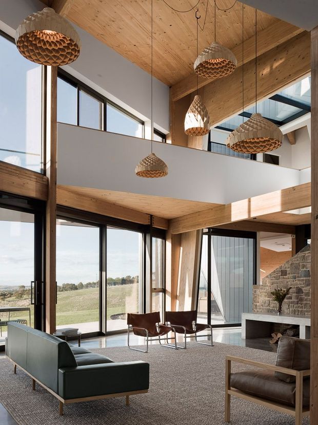 A spacious double-height living room with a mezzanine overlooking the space. The room features extensive floor-to-ceiling glazing, exposed timber beams and ceilings, a large stone fireplace, suspended sculptural timber pendant lights, and contemporary leather seating arranged around a low coffee table. The interior opens to expansive rural views and is filled with natural light.