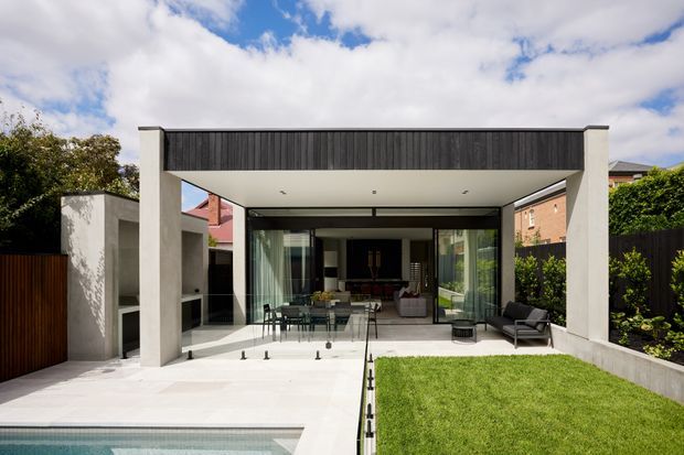 A contemporary rear exterior of a suburban home featuring a deep covered outdoor entertaining terrace framed by concrete columns, dark vertical cladding, and extensive floor-to-ceiling glazing. The terrace opens to a dining and living area, with pale paved flooring, a lawn, landscaped boundary planting, and the edge of a swimming pool in the foreground.