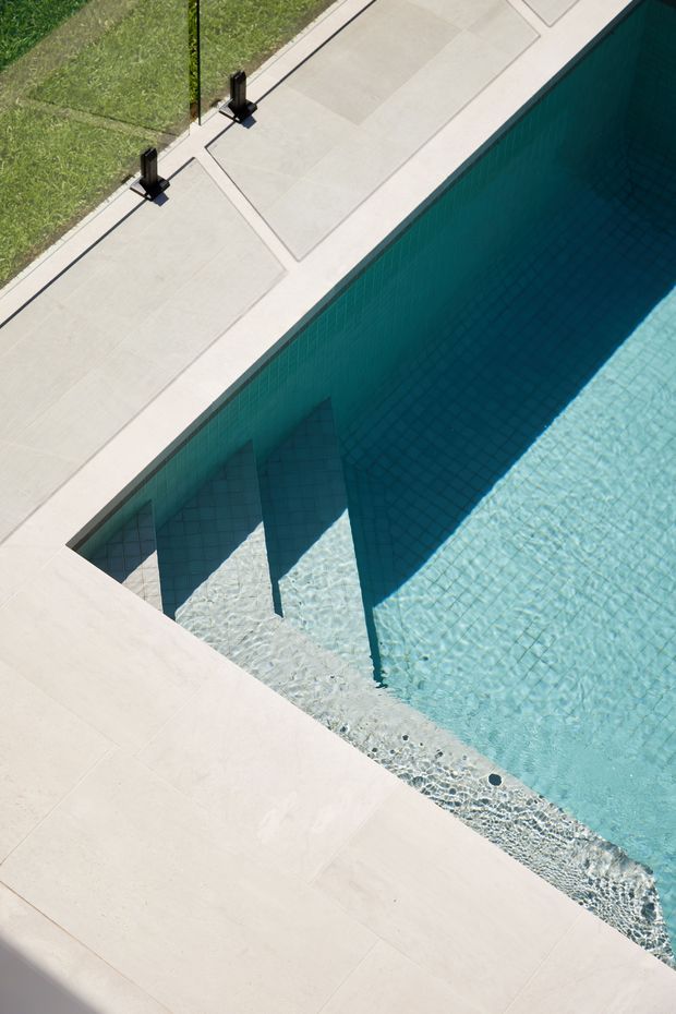 High-angle close-up of a modern swimming pool with clear turquoise water, pale rectangular poolside paving, dark pool edging, and a narrow strip of lawn. Strong geometric lines and architectural shadows create a clean, contemporary outdoor composition.