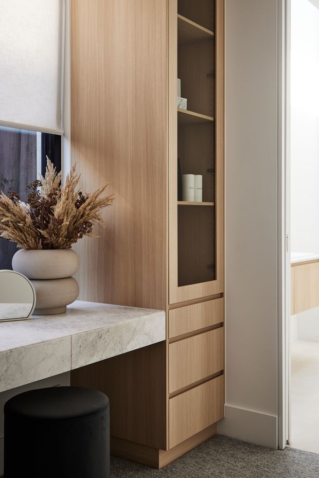 A refined built-in dressing or bedroom storage area featuring warm light-wood cabinetry, open shelving, drawers, and a pale natural-stone vanity ledge. A black upholstered stool, rounded ceramic vessel with dried botanicals, and small mirror sit beside a window with a roller blind. An open doorway reveals part of an adjacent light-toned room. The design is clean, warm, and contemporary with integrated joinery and soft natural textures.