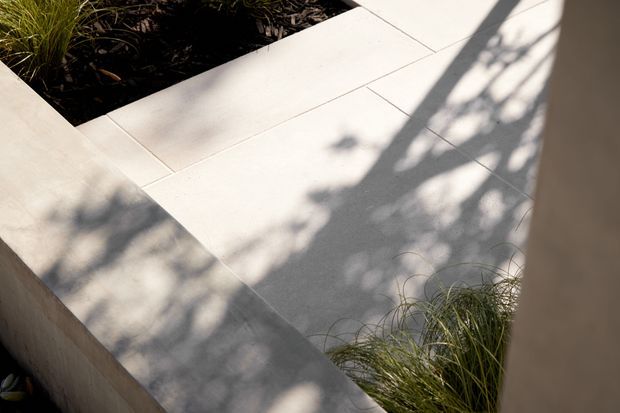 Close-up view of a pale tiled outdoor courtyard or terrace, with broad rectangular paving slabs, low rendered or concrete edges, planted garden beds, and soft shadows cast by surrounding vegetation. The image focuses on the landscape paving and built garden geometry rather than a building interior.