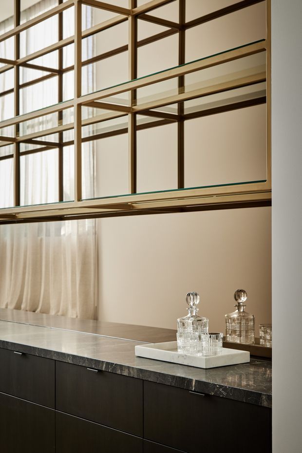 A refined interior joinery detail featuring a long dark built-in cabinet with a veined stone benchtop, topped by an integrated mirrored and glass shelving display framed in warm bronze-toned metal. Crystal decanters and tumblers sit on a pale tray, while sheer curtains and soft beige walls create a warm, elegant atmosphere. The image focuses on the fixed cabinetry, display shelving and reflective surfaces rather than a complete room.