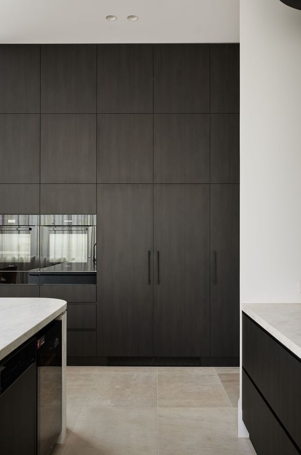Contemporary kitchen interior with a tall wall of dark charcoal wood-grain cabinetry, integrated black appliances, pale stone benchtops, and large light beige floor tiles. A compact island is visible in the foreground, with a reflective splashback or window area behind it. The room has a restrained, high-contrast palette and clean, handle-minimal joinery.
