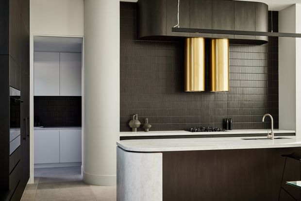 Sophisticated contemporary kitchen with a large island featuring a curved pale stone end panel and dark wood cabinetry. The kitchen has a dark vertically textured tile splashback, integrated appliances, a gas cooktop, undermount sink with a curved metal tap, and a dramatic suspended dark canopy with twin cylindrical brass-gold pendant lights. A tall rounded structural column separates the main kitchen from a secondary service or pantry area with pale cabinetry.